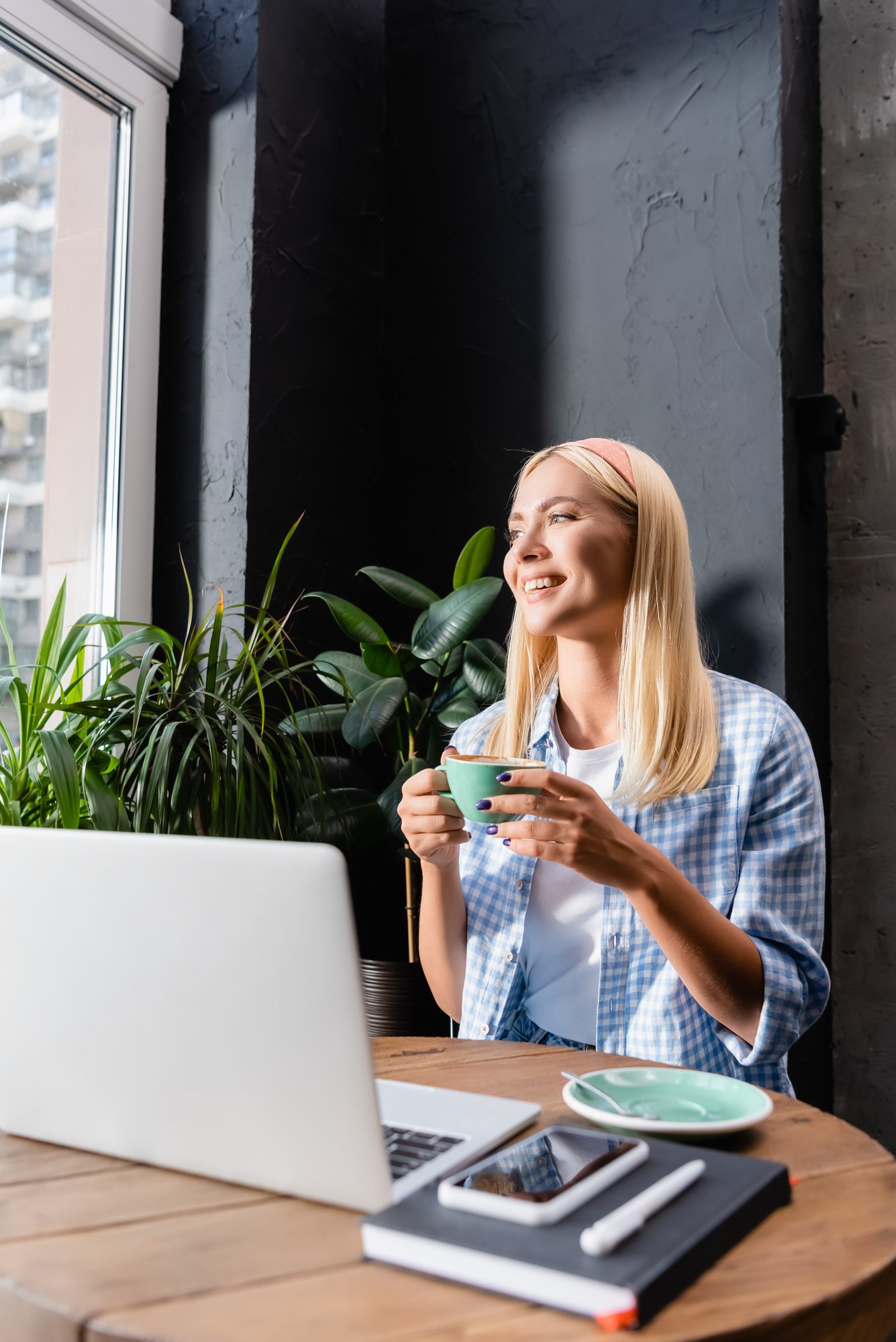 Happy freelancer holding cup of coffee near laptop on blurred foreground