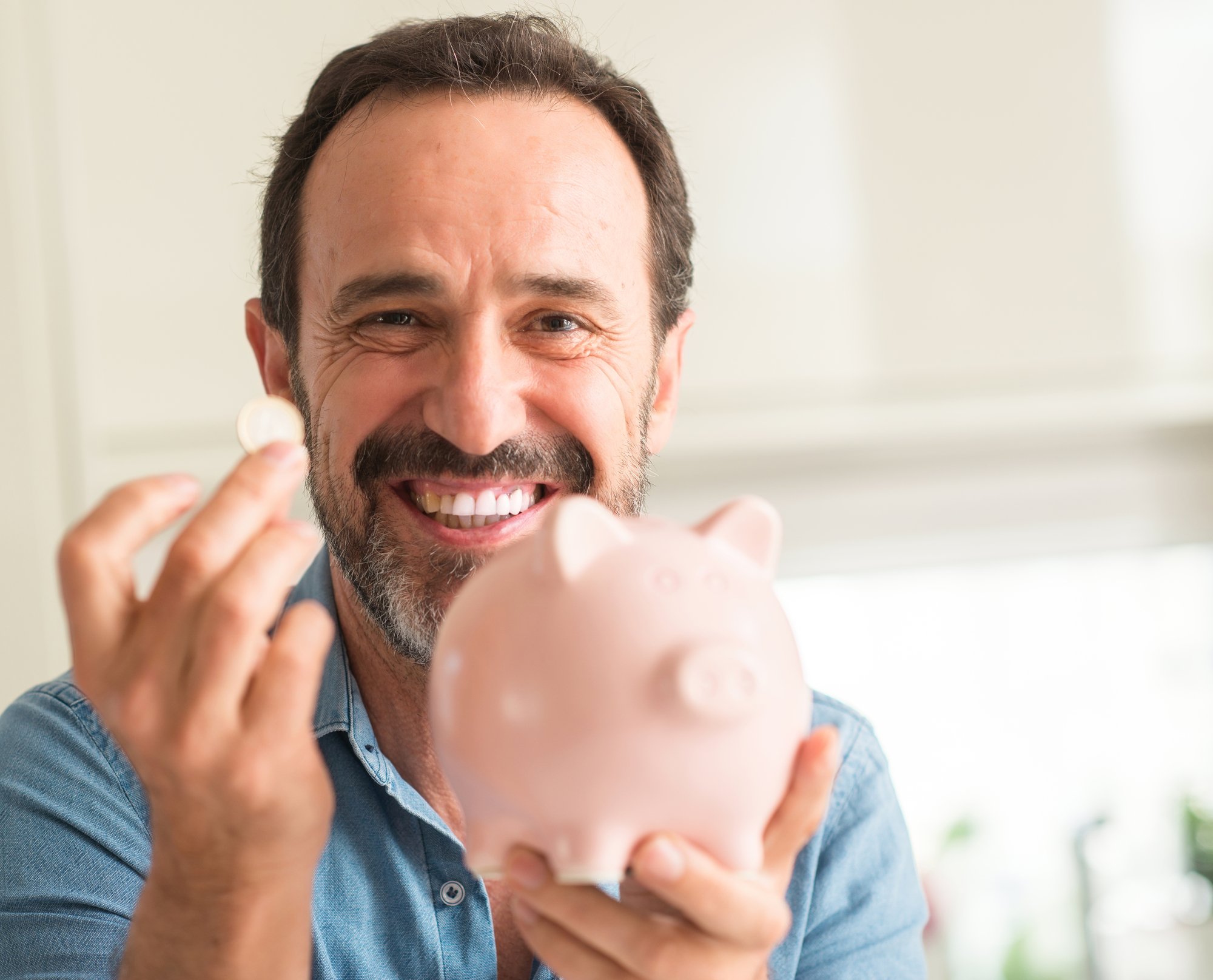 Middle age man save money on piggy bank with a happy face standing and smiling with a confident smile showing teeth