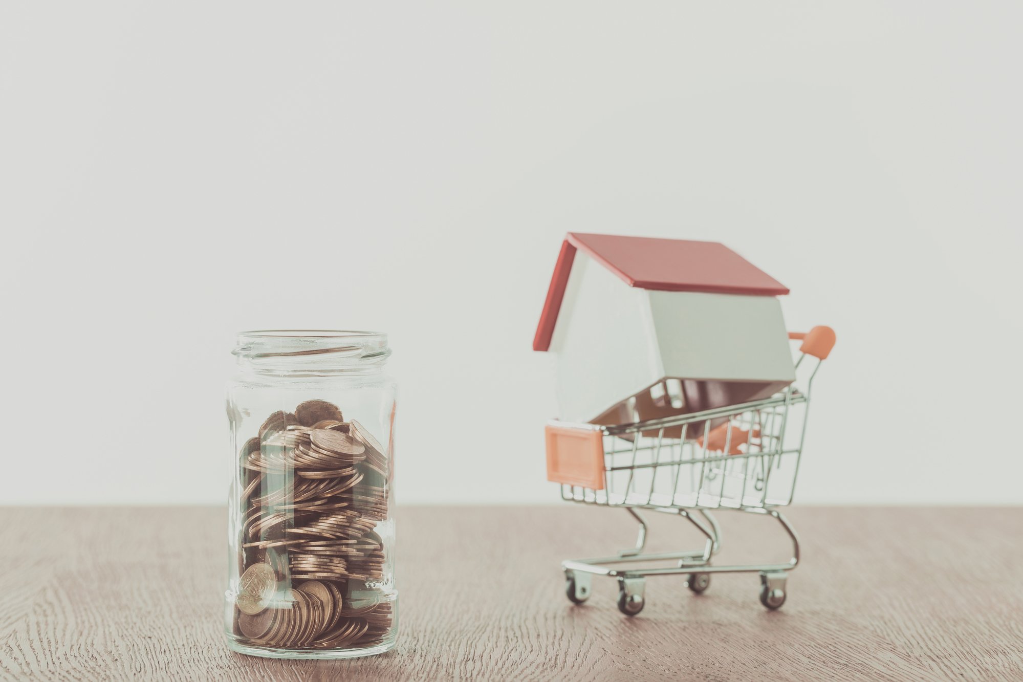 Small wooden house on supermarket cart, jar of coins on wooden table, saving concept