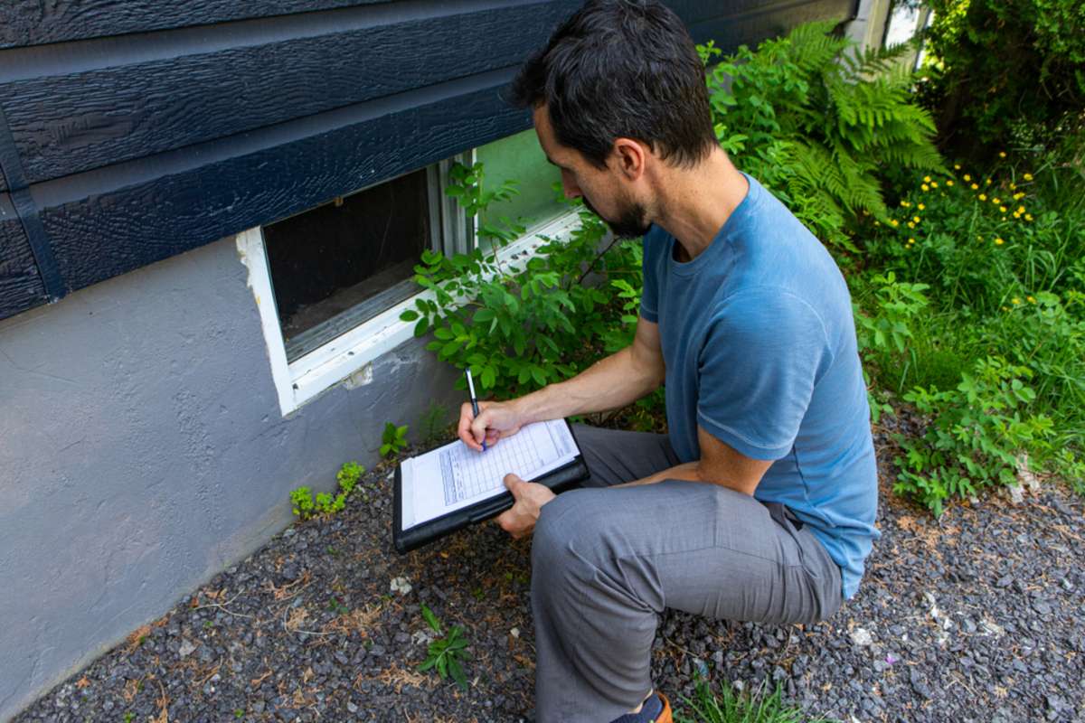 A close up view on environmental home quality inspector at work, filling in a form as he inspects the exterior of a cellar window