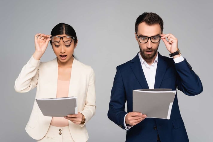 Surprised interracial business partners looking at documents and touching eyeglasses isolated on grey