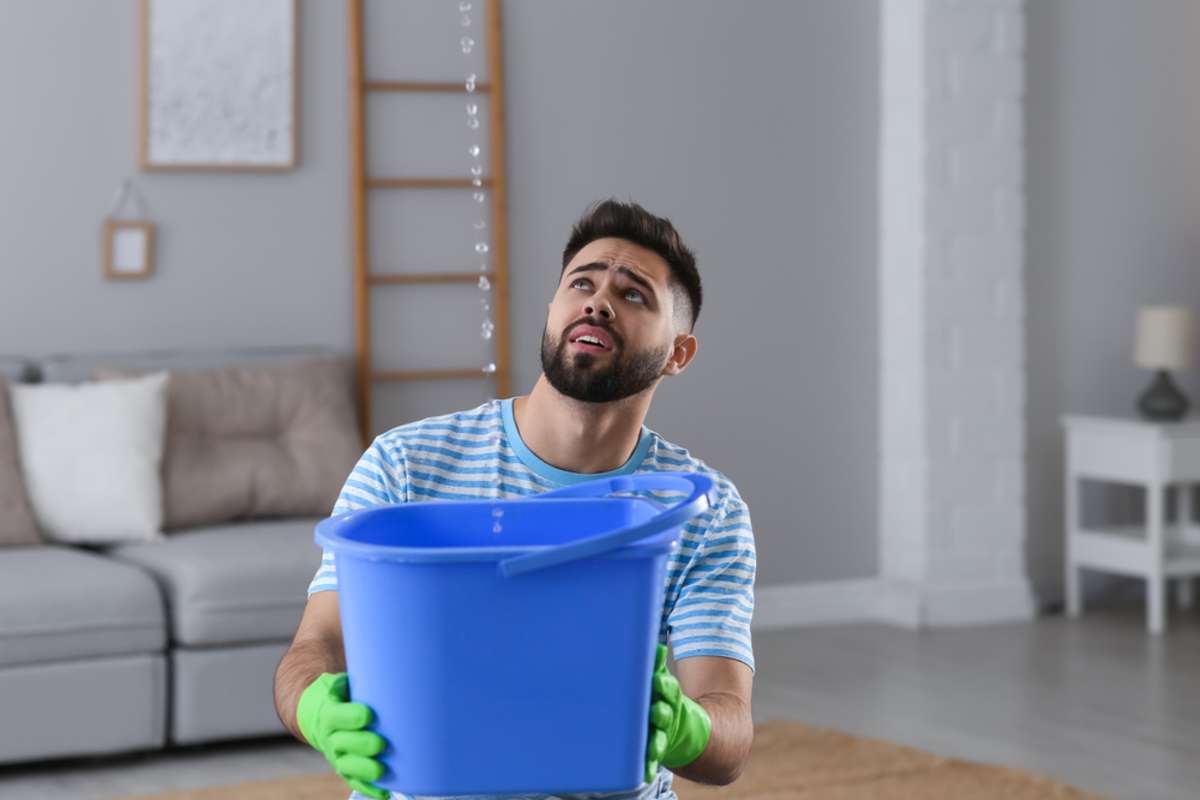 Young man collecting leaking water from ceiling at home, space for text