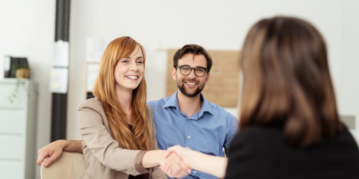 Happy young couple meeting with a broker in her office leaning over the desk to shake hands